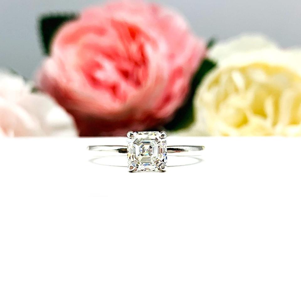 Sterling silver 4-prong asscher cut DEF moissanite solitaire engagement ring. The ring is resting on a white surface with a background of pink and white flowers.