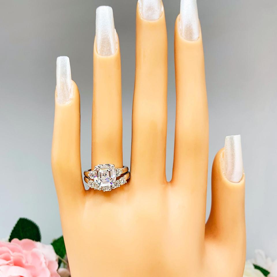 Mannequin hand with long white shimmery nails, displayed against a soft white-gray background with pink and white floral accents. The hand is wearing a sterling silver 5-stone curved wedding band featuring 3mm round precision faceted moissanite stones in prong settings. The ring is paired with an asscher with round side stone 3-stone cz engagement ring. This ring is not included.