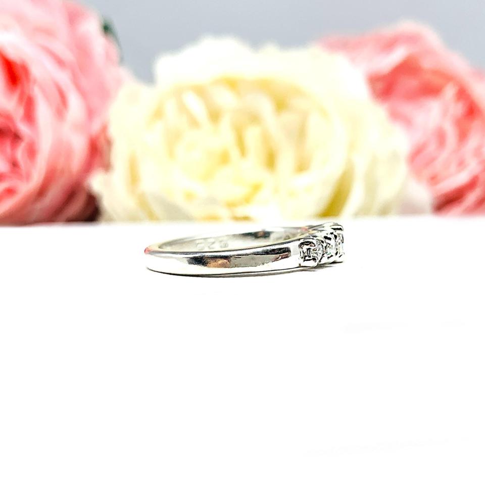 A sterling silver graduated moissanite engagement ring featuring a 4mm center stone flanked by 3mm and 2.5mm side stones, shown in a close-up against a soft-focus background of pink and white flowers. The moissanites sparkle with icy brilliance, making this an elegant alternative engagement or anniversary ring.