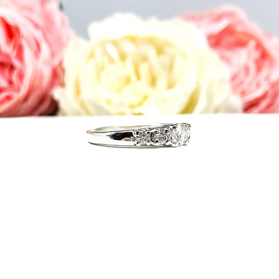 A sterling silver graduated moissanite engagement ring featuring a 4mm center stone flanked by 3mm and 2.5mm side stones, shown in a close-up against a soft-focus background of pink and white flowers. The moissanites sparkle with icy brilliance, making this an elegant alternative engagement or anniversary ring.