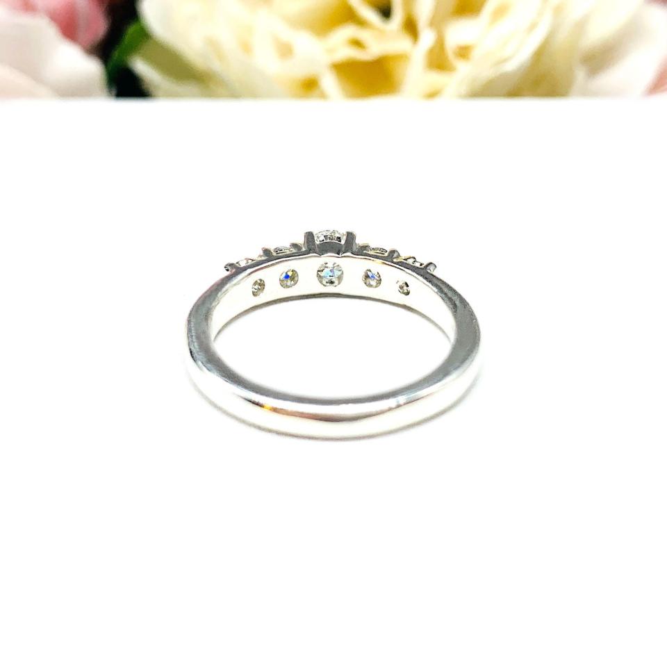 A sterling silver graduated moissanite engagement ring featuring a 4mm center stone flanked by 3mm and 2.5mm side stones, shown in a close-up against a soft-focus background of pink and white flowers. The moissanites sparkle with icy brilliance, making this an elegant alternative engagement or anniversary ring.