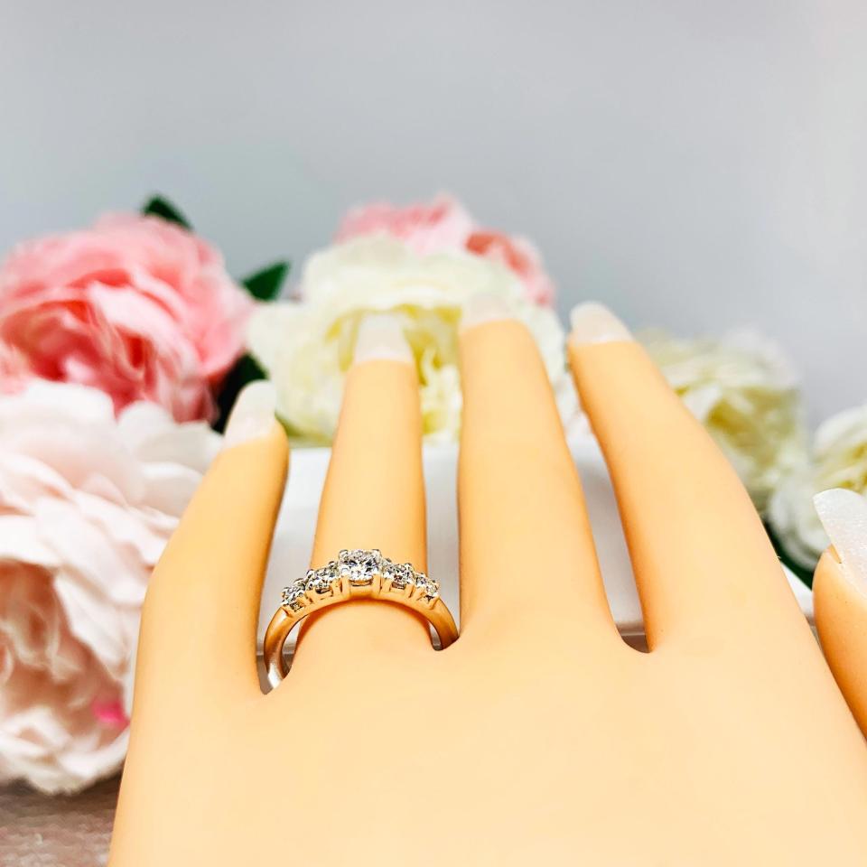 Mannequin hand with long white shimmery nails, displayed against a soft white-gray background with pink and white floral accents. The hand is wearing  a sterling silver graduated moissanite engagement ring featuring a 4mm center stone flanked by 3mm and 2.5mm side stones.
