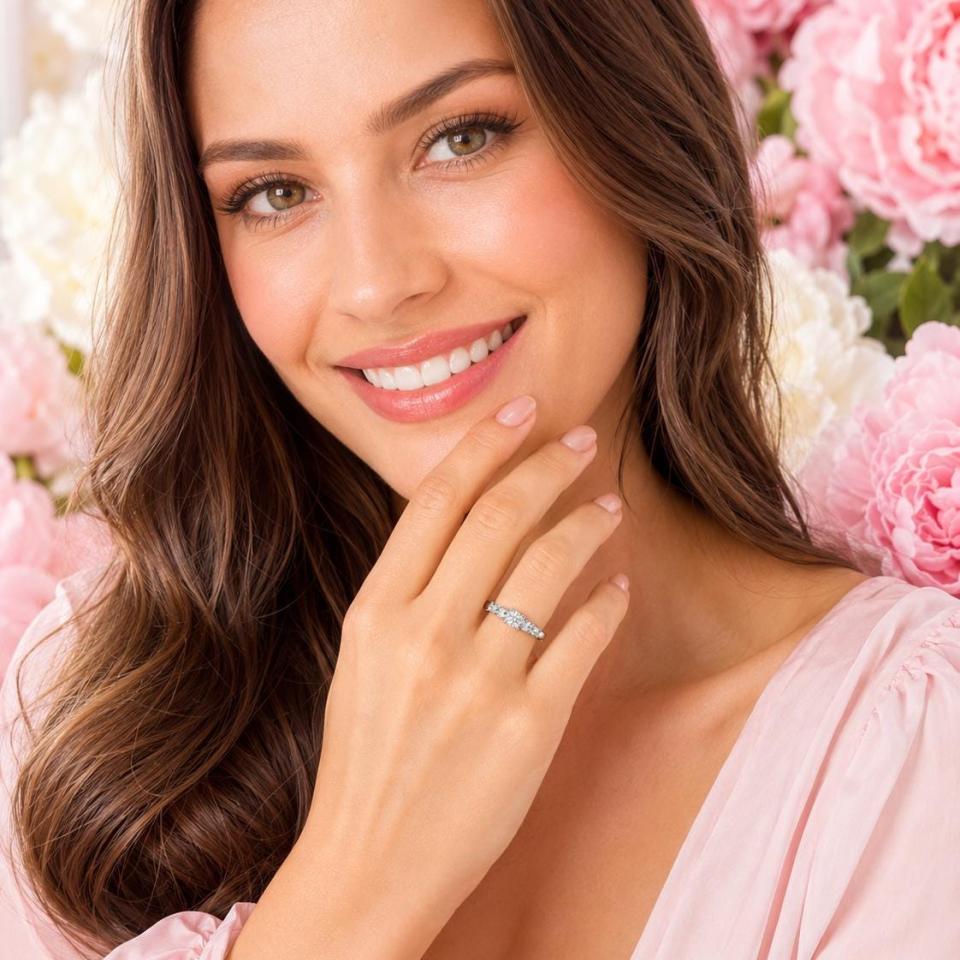 A sterling silver graduated moissanite engagement ring featuring a 4mm center stone flanked by 3mm and 2.5mm side stones, shown on a woman against a backdrop of pink and white flowers. Model image for size and styling reference — not a photo of the actual ring.