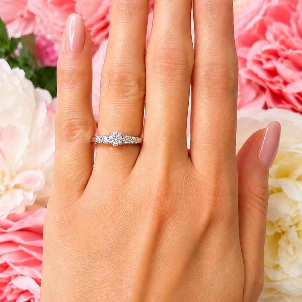 A sterling silver graduated moissanite engagement ring featuring a 4mm center stone flanked by 3mm and 2.5mm side stones, shown on a hand with soft pink manicured nails against a backdrop of pink and white flowers. Model image for size and styling reference — not a photo of the actual ring.