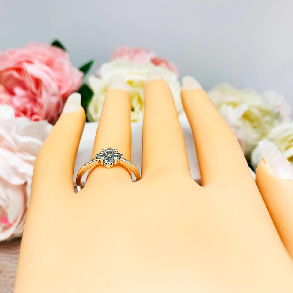 Mannequin hand with long white shimmery nails, displayed against a soft white-gray background with pink and white floral accents. The hand is wearing a sterling silver  cluster ring featuring four 3mm round light blue aquamarine gemstones.