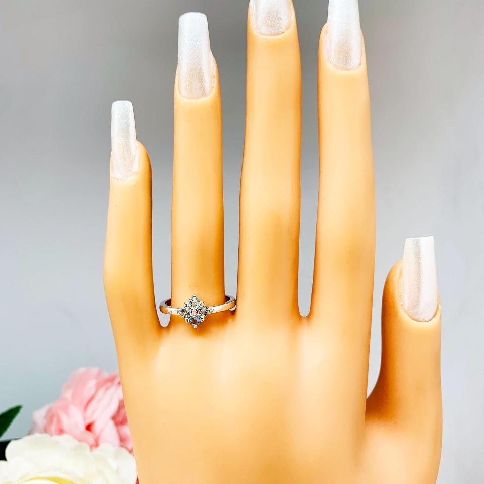 Mannequin hand with long white shimmery nails, displayed against a soft white-gray background with pink and white floral accents. The hand is wearing a sterling silver  cluster ring featuring four 3mm round light blue aquamarine gemstones.