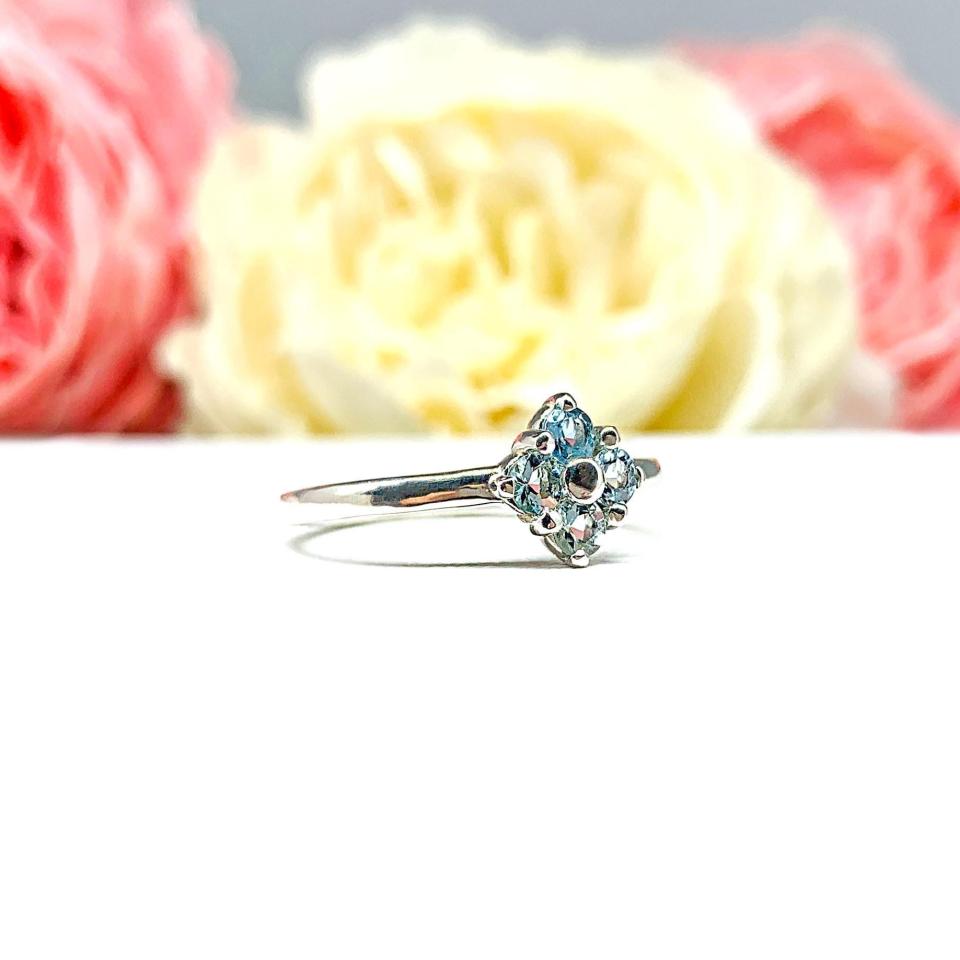 Sterling silver cluster ring featuring four 3mm round light blue aquamarine gemstones. Displayed against a soft-focus floral background, the ring sparkles with contrast and elegance.