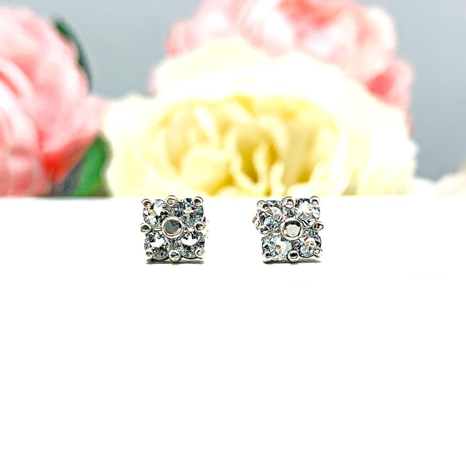 Sterling silver cluster earrings featuring four 3mm round light blue aquamarine gemstones. Displayed against a soft-focus floral background, the earrings sparkle with contrast and elegance.