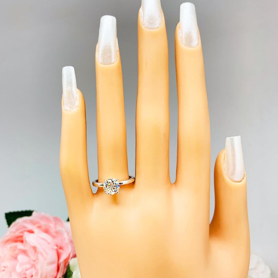 Mannequin hand with long white shimmery nails, displayed against a soft white-gray background with pink and white floral accents. The hand is wearing a solitaire engagement ring with a round brilliant light champagne moissanite. The setting is a classic four-prong design, and the band is a simple, sterling silver band.
