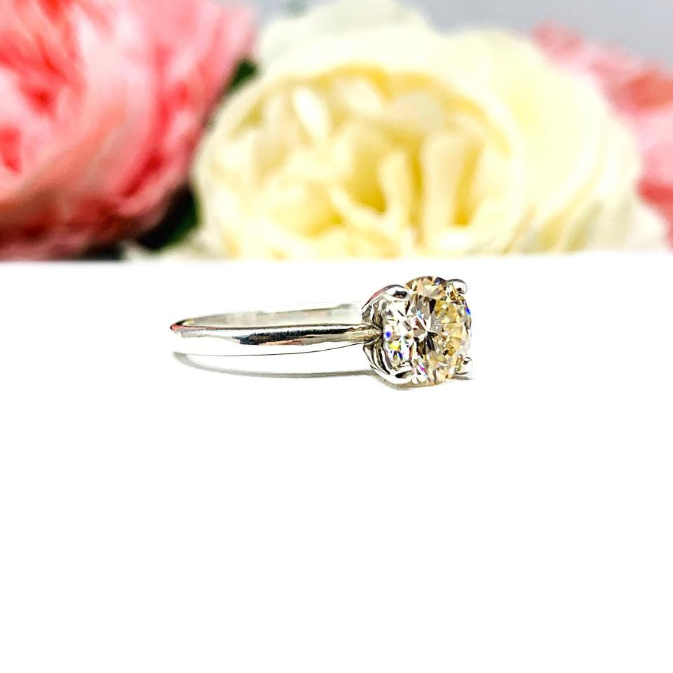 A solitaire engagement ring with a round brilliant light champagne moissanite. The setting is a classic four-prong design, and the band is a simple, sterling silver band. The ring is displayed on a white background with blurred pink and yellow flowers in the background.