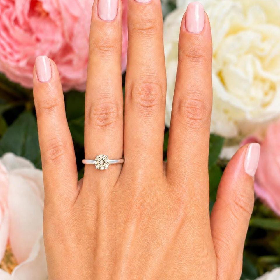 Woman's hand wearing a sterling silver 6.5mm round-cut 4-prong light champagne moissanite engagement ring. Model image for size and styling reference — not a photo of the actual ring.