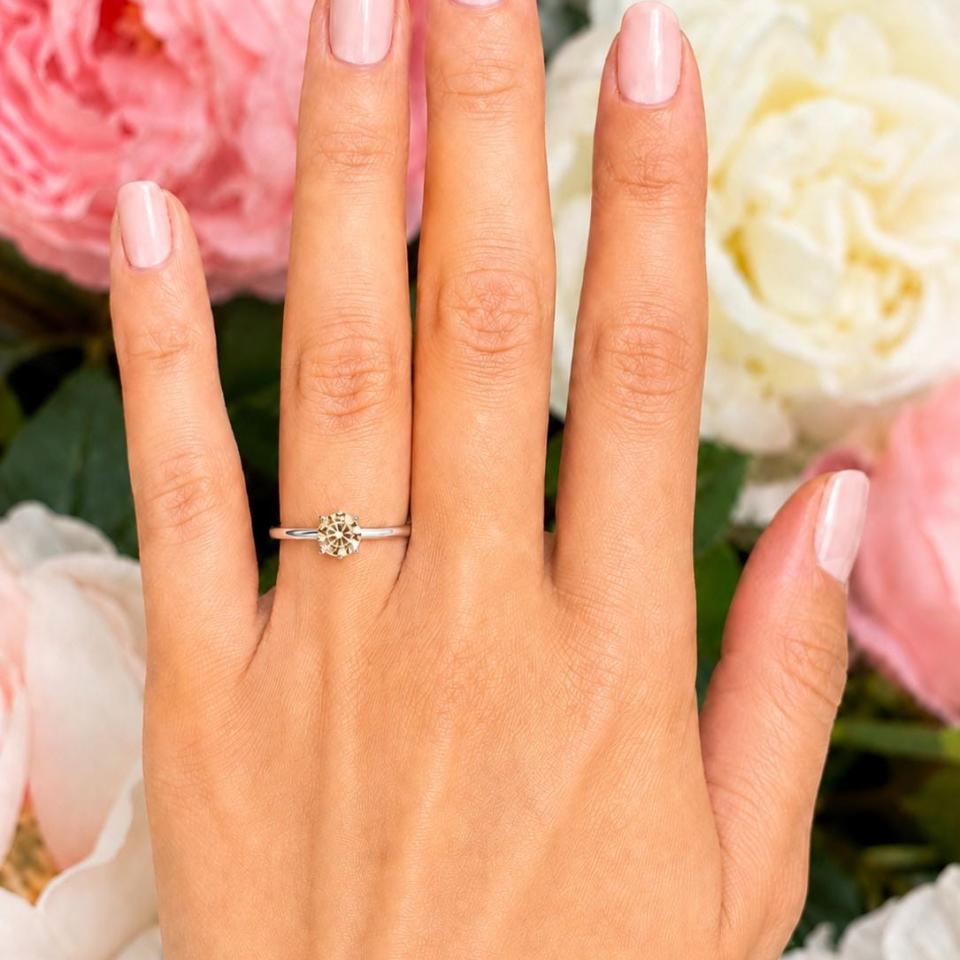 Woman's hand wearing a sterling silver 5mm round-cut 4-prong light champagne moissanite engagement ring. Model image for size and styling reference — not a photo of the actual ring.