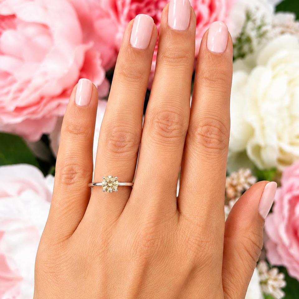 Woman's hand wearing a sterling silver 7mm round-cut 4-prong light champagne moissanite engagement ring. Model image for size and styling reference — not a photo of the actual ring.