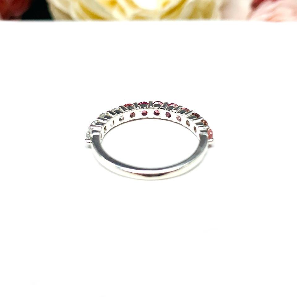 Sterling silver twelve stone band set with 2.5mm round gemstones in this configuration: four white topaz, four pink tourmaline and four light pink tourmaline set in a shared prong setting, displayed on a white surface with soft-focus pastel flowers in the background.