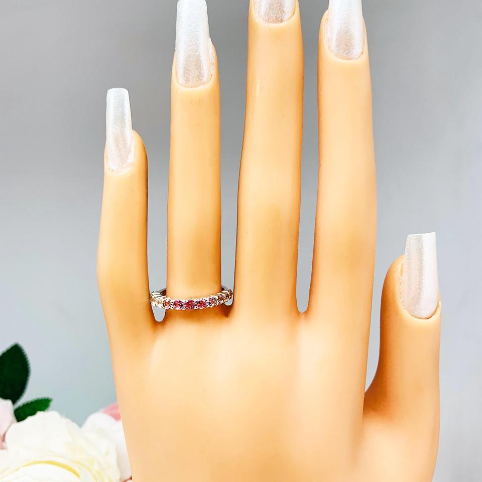 Mannequin hand with long white shimmery nails, displayed against a soft white-gray background with pink and white floral accents. The hand is wearing a sterling silver  twelve stone band set with 2.5mm round gemstones in this configuration: four white topaz, four pink tourmaline and four light pink tourmaline set in a shared prong setting.