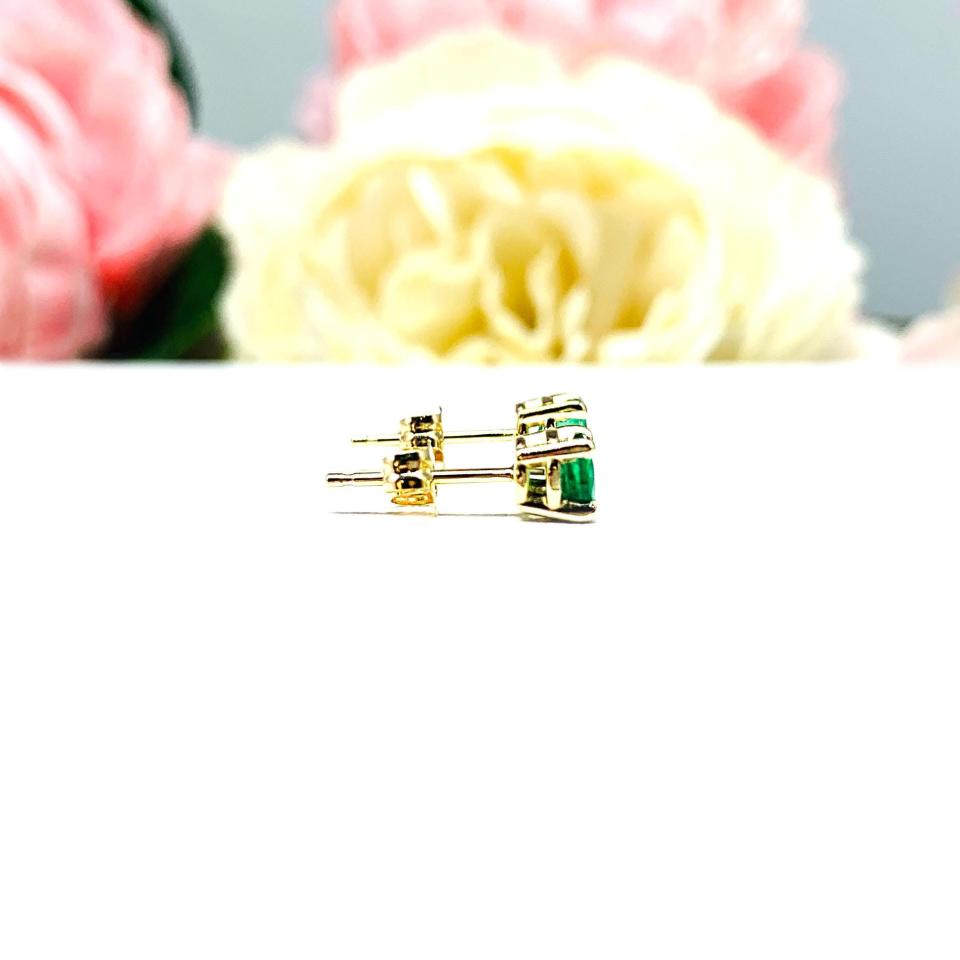 A pair of 14k yellow gold stud earrings set with 4mm round natural emerald gemstones. The four prong basket earrings are on a white surface with both earrings facing to the right to showcase the center post and butterfly friction clasps.