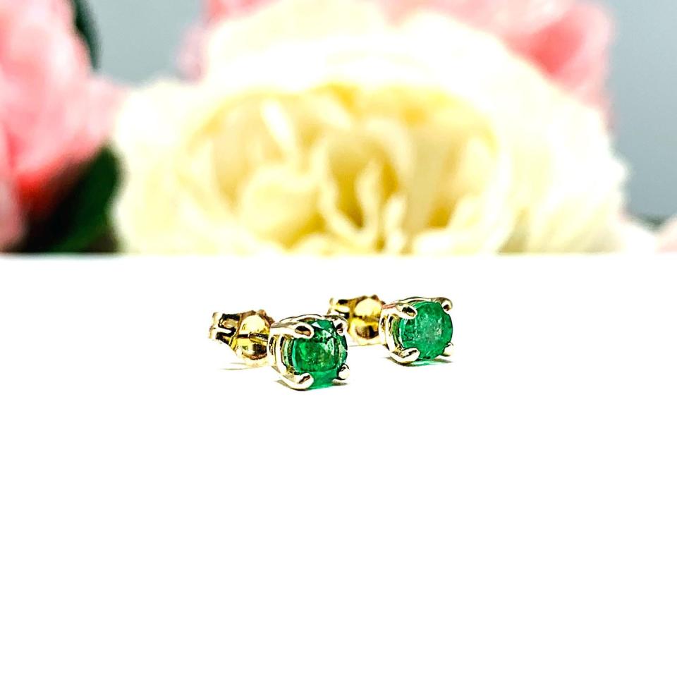 A pair of 14k yellow gold stud earrings set with 4mm round natural emerald gemstones. The four prong basket earrings are on a white surface with both earrings facing slightly to the right to showcase the clarity and vibrant green color of the stones and to display the center post and butterfly friction clasps.