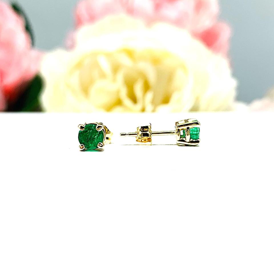 A pair of 14k yellow gold stud earrings set with 4mm round natural emerald gemstones. The four prong basket earrings are on a white surface with the left earring facing front to showcase the clarity and vibrant green color of the stones, and the right earring facing toward the right to display the center post and butterfly friction clasp.