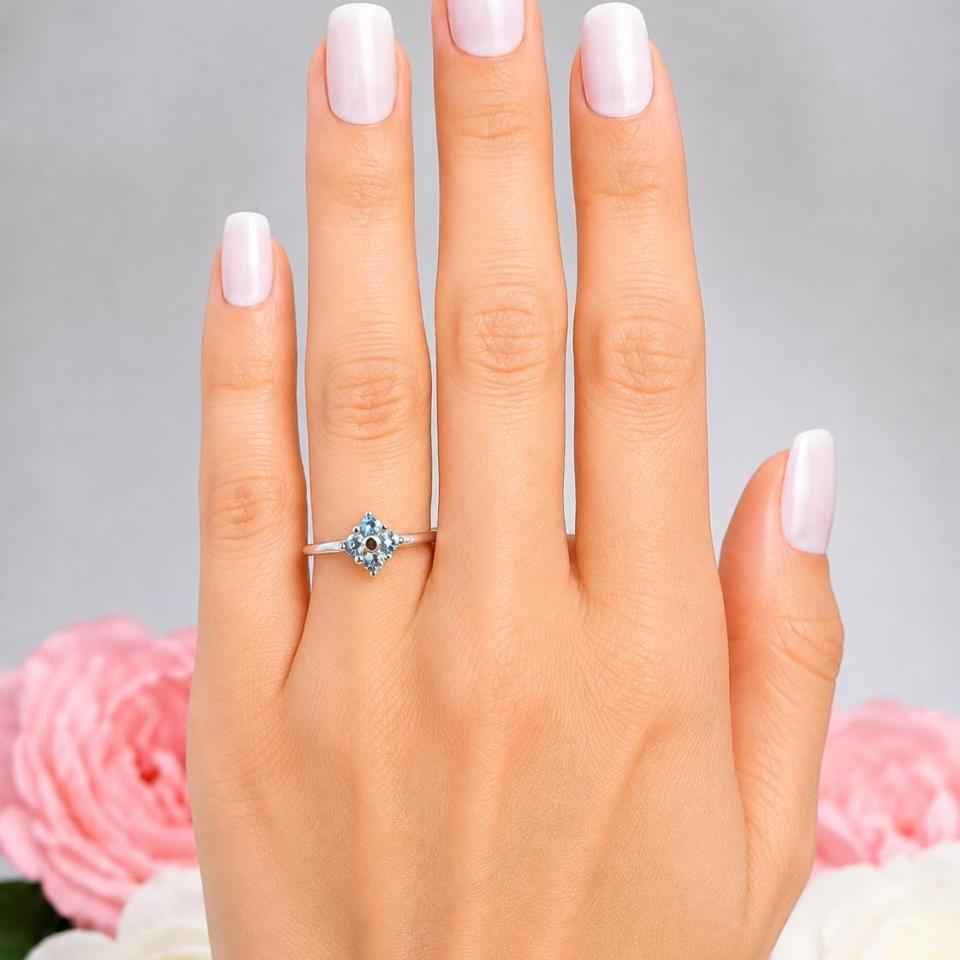 A woman's hand wearing a sterling silver cluster ring featuring four 3mm round light blue aquamarine gemstones. Displayed against a soft-focus floral background, the ring sparkles with contrast and elegance. Model image for size and styling reference — not a photo of the actual ring.