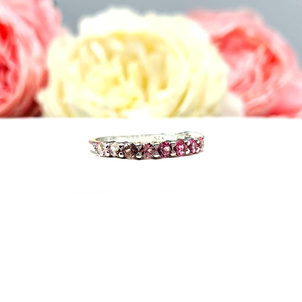 Sterling silver twelve stone band set with 2.5mm round gemstones in this configuration: four white topaz, four pink tourmaline and four light pink tourmaline set in a shared prong setting, displayed on a white surface with soft-focus pastel flowers in the background.