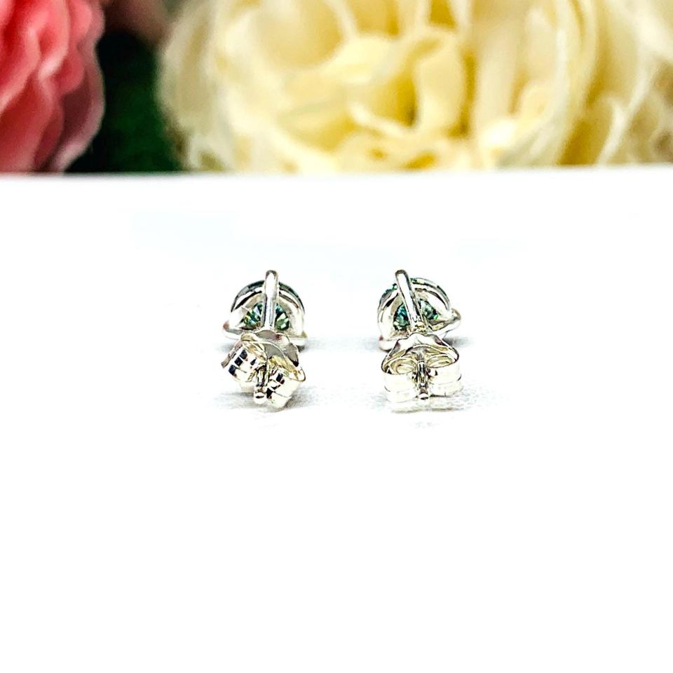 Sterling silver stud earrings featuring round authentic sea foam green moissanite stones in martini-style settings, displayed on a white surface with soft floral elements in the background.