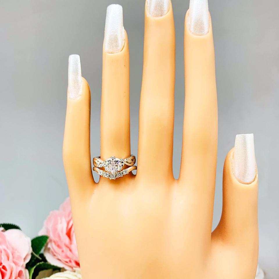 Mannequin hand with long white shimmery nails, displayed against a soft white-gray background with pink and white floral accents. The hand is wearing a sterling silver 5-stone curved wedding band featuring 2mm round precision faceted moissanite stones in prong settings. Shown with a round moissanite solitiare with leaf design (not included).