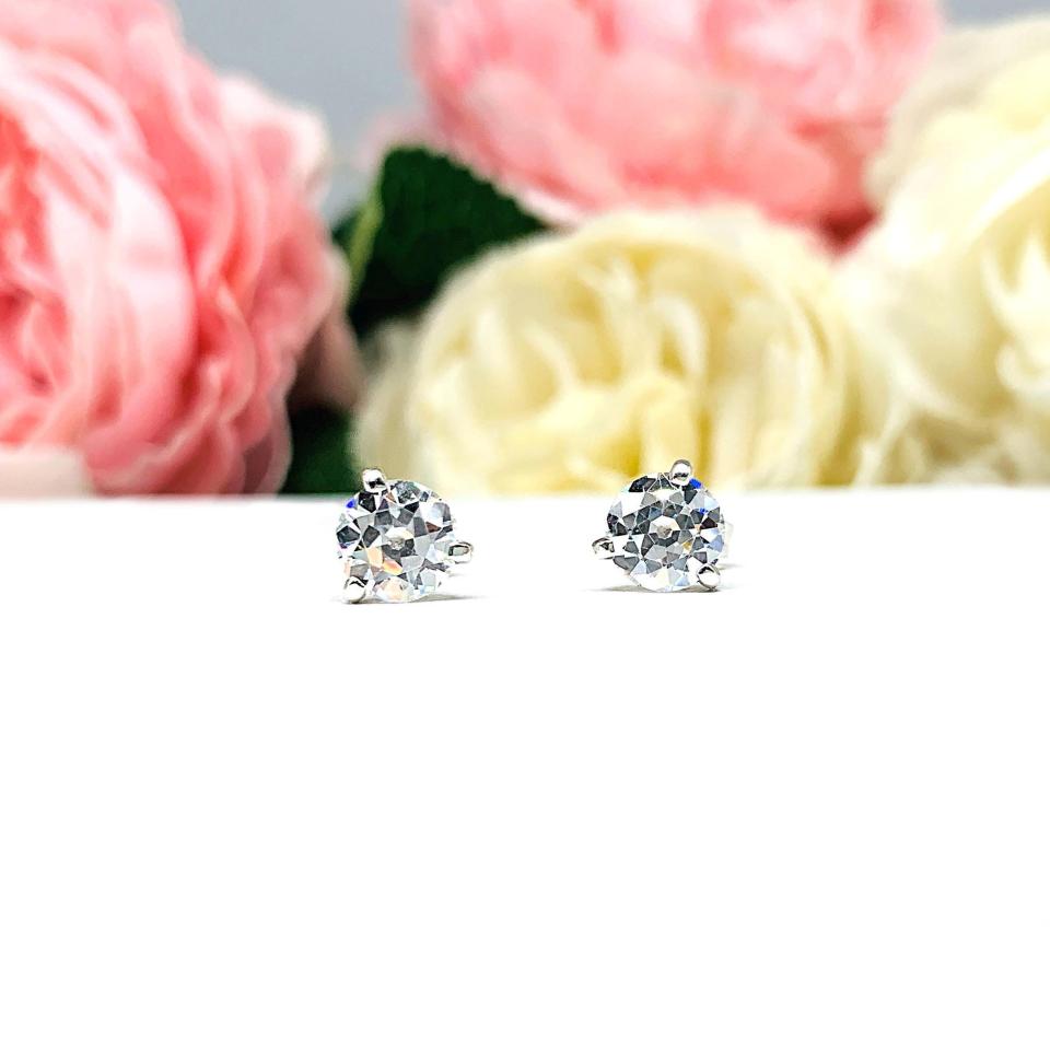 A pair of sterling silver 3-prong martini stud earrings with 6mm colorless old European cut cubic zirconias. The earrings are resting on a white surface with a background of pink and white flowers.