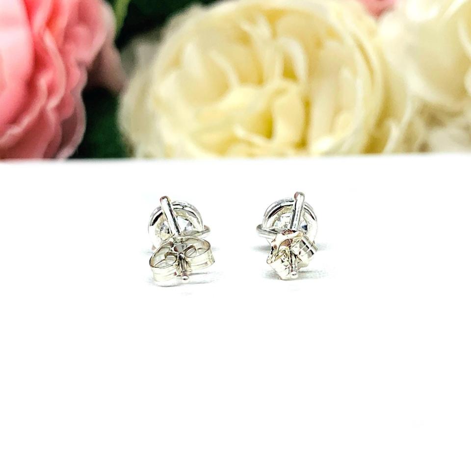 A pair of sterling silver 3-prong martini stud earrings with 6mm colorless old European cut cubic zirconias. The earrings are resting on a white surface with a background of pink and white flowers.