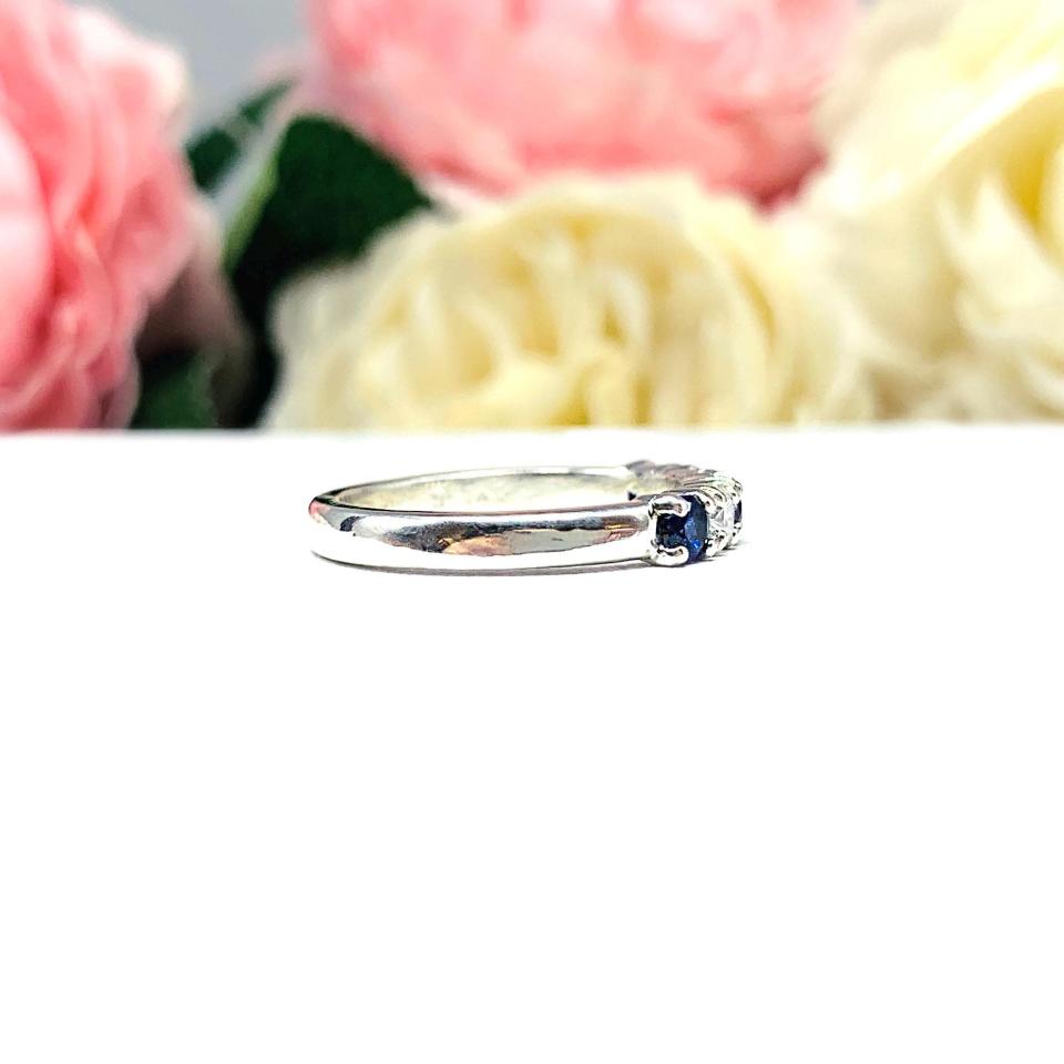 A sterling silver 5-stone band featuring alternating round lab blue sapphires and moissanites. The band has a classic design with the gemstones set in a row with three sapphires and two moissanites. Displayed on a white surface with a pastel floral background.