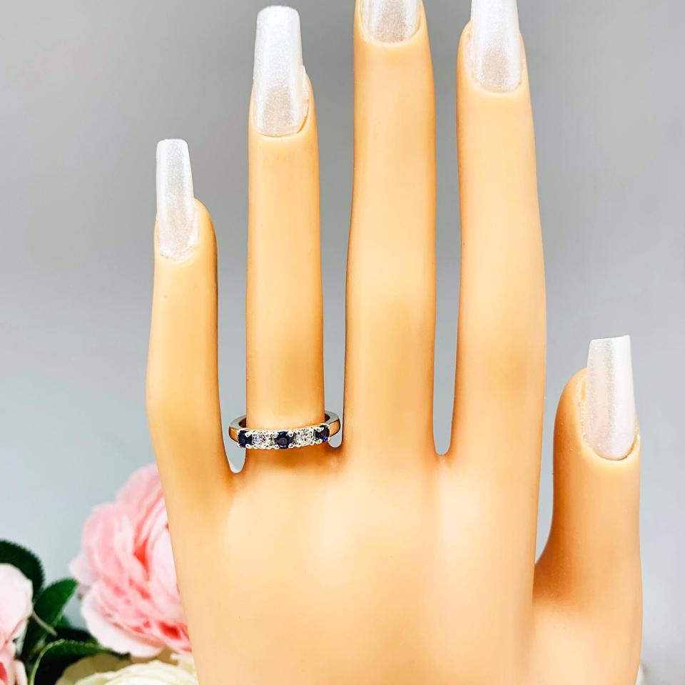 Mannequin hand with long white shimmery nails, displayed against a soft white-gray background with pink and white floral accents. The hand is wearing a sterling silver 5-stone band featuring alternating round lab blue sapphires and moissanites. The band has a classic design with the gemstones set in a row with three sapphires and two moissanites.