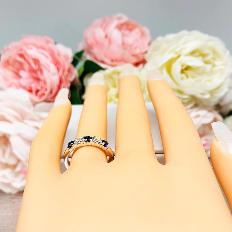 Mannequin hand with long white shimmery nails, displayed against a soft white-gray background with pink and white floral accents. The hand is wearing a sterling silver 5-stone band featuring alternating round lab blue sapphires and moissanites. The band has a classic design with the gemstones set in a row with three sapphires and two moissanites.