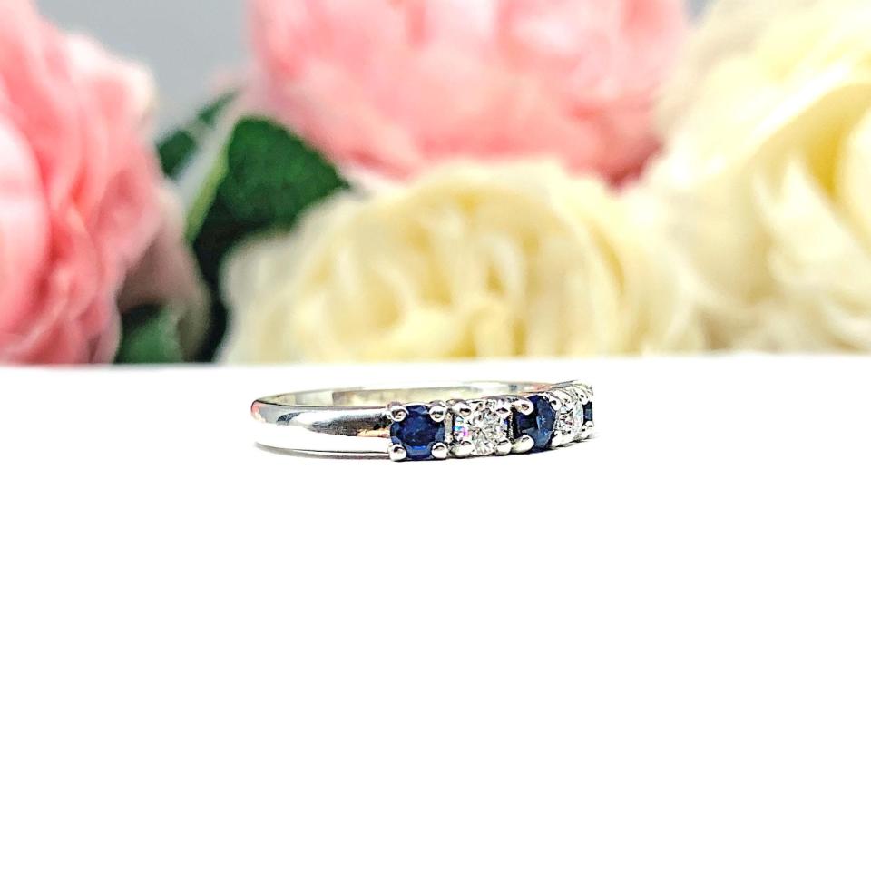 A sterling silver 5-stone band featuring alternating round lab blue sapphires and moissanites. The band has a classic design with the gemstones set in a row with three sapphires and two moissanites. Displayed on a white surface with a pastel floral background.