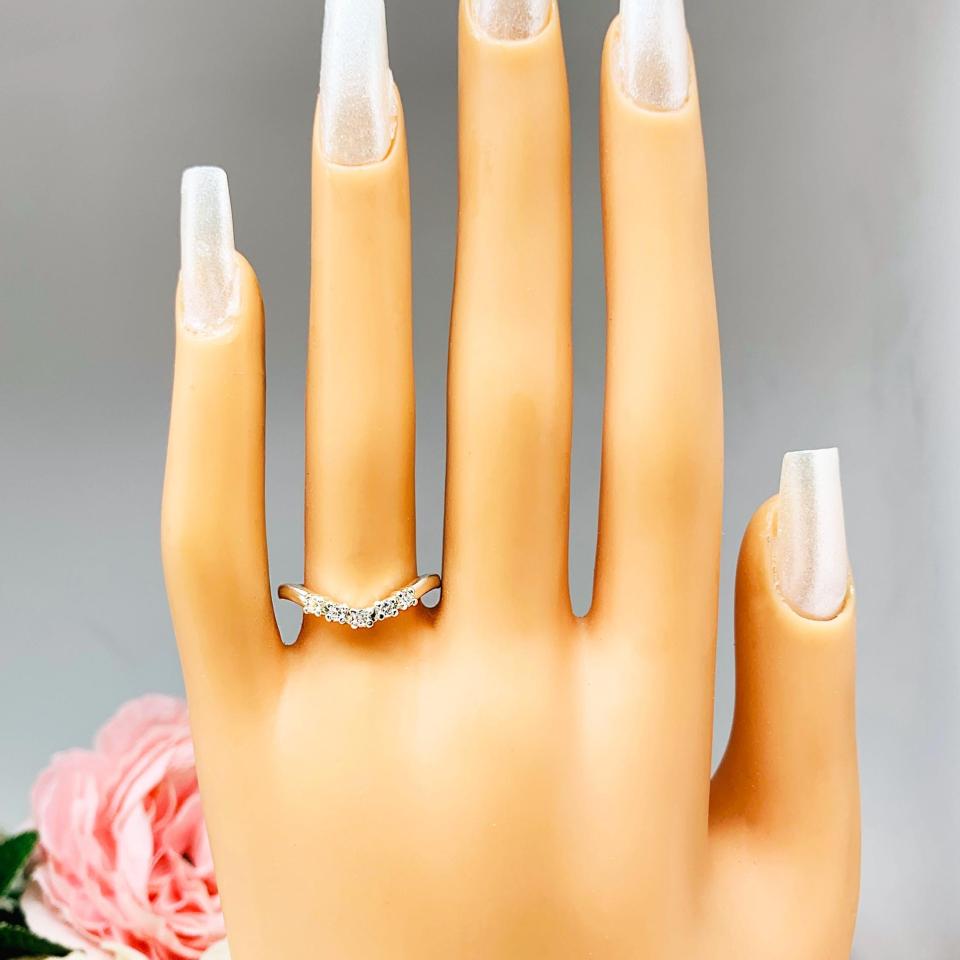 Mannequin hand with long white shimmery nails, displayed against a soft white-gray background with pink and white floral accents. The hand is wearing a sterling silver 5-stone curved wedding band featuring 2mm round precision faceted moissanite stones in prong settings.