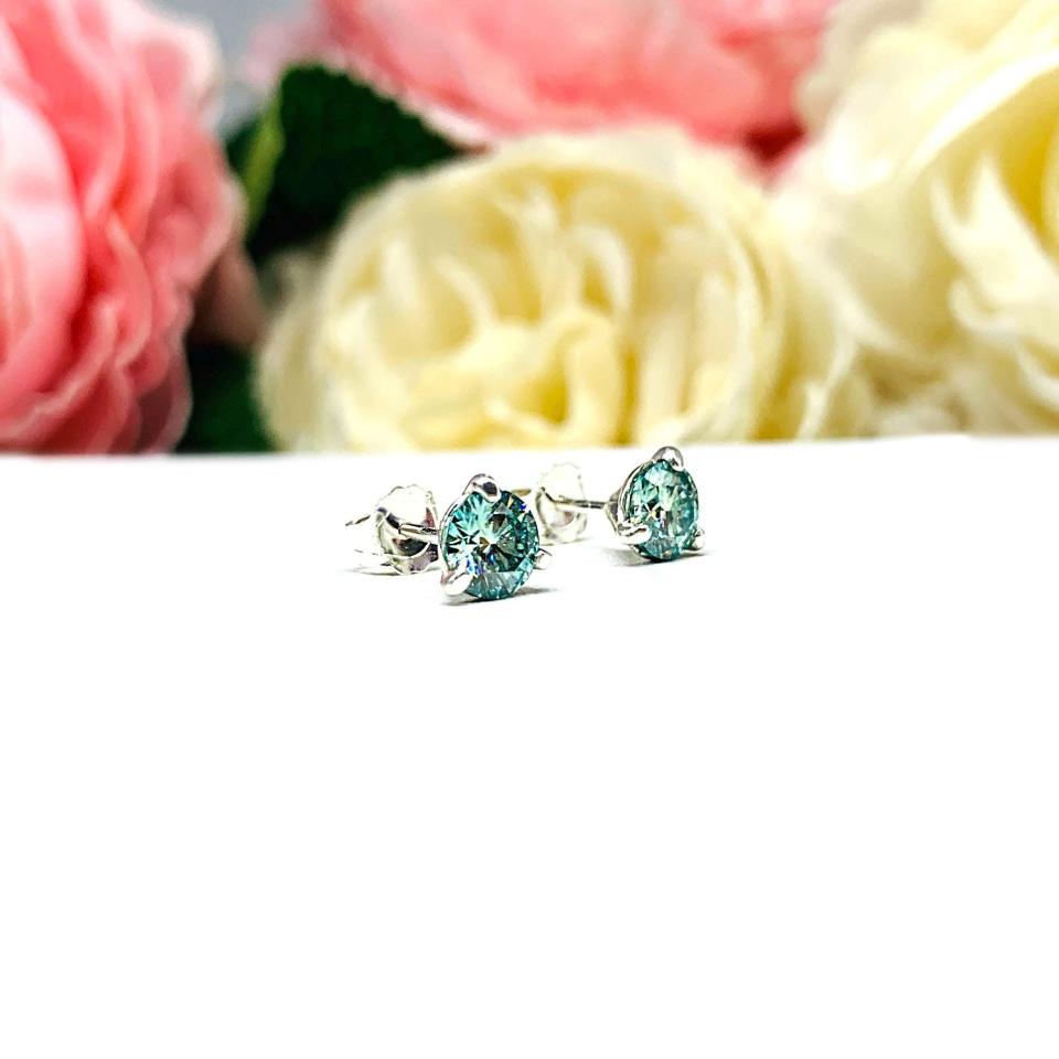 Sterling silver stud earrings featuring round authentic sea foam green moissanite stones in martini-style settings, displayed on a white surface with soft floral elements in the background.