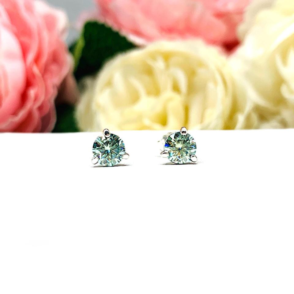 Sterling silver stud earrings featuring round authentic sea foam green moissanite stones in martini-style settings, displayed on a white surface with soft floral elements in the background.