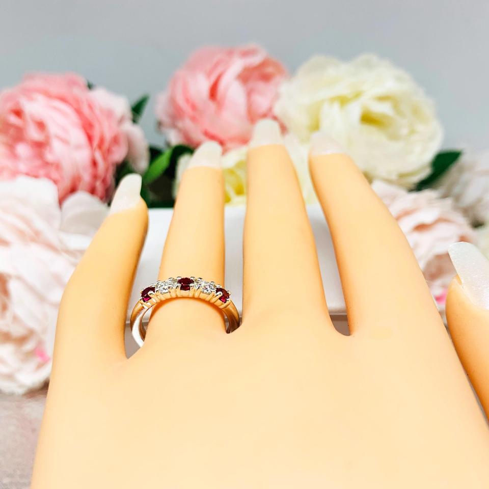 Mannequin hand with long white shimmery nails, displayed against a soft white-gray background with pink and white floral accents. The hand is wearing a sterling silver 5-stone band featuring alternating round lab rubies and moissanites. The band has a classic design with the gemstones set in a row with three rubies and two moissanites.