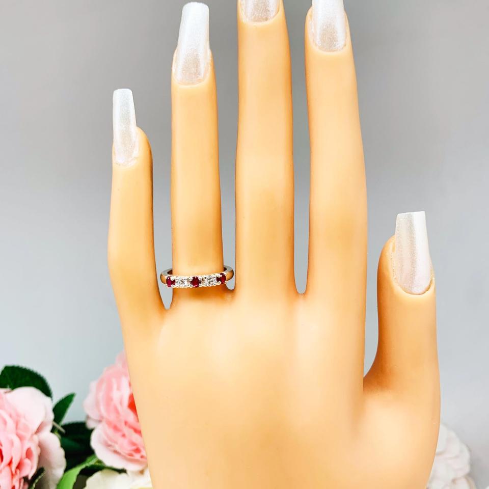 Mannequin hand with long white shimmery nails, displayed against a soft white-gray background with pink and white floral accents. The hand is wearing a sterling silver 5-stone band featuring alternating round lab rubies and moissanites. The band has a classic design with the gemstones set in a row with three rubies and two moissanites.