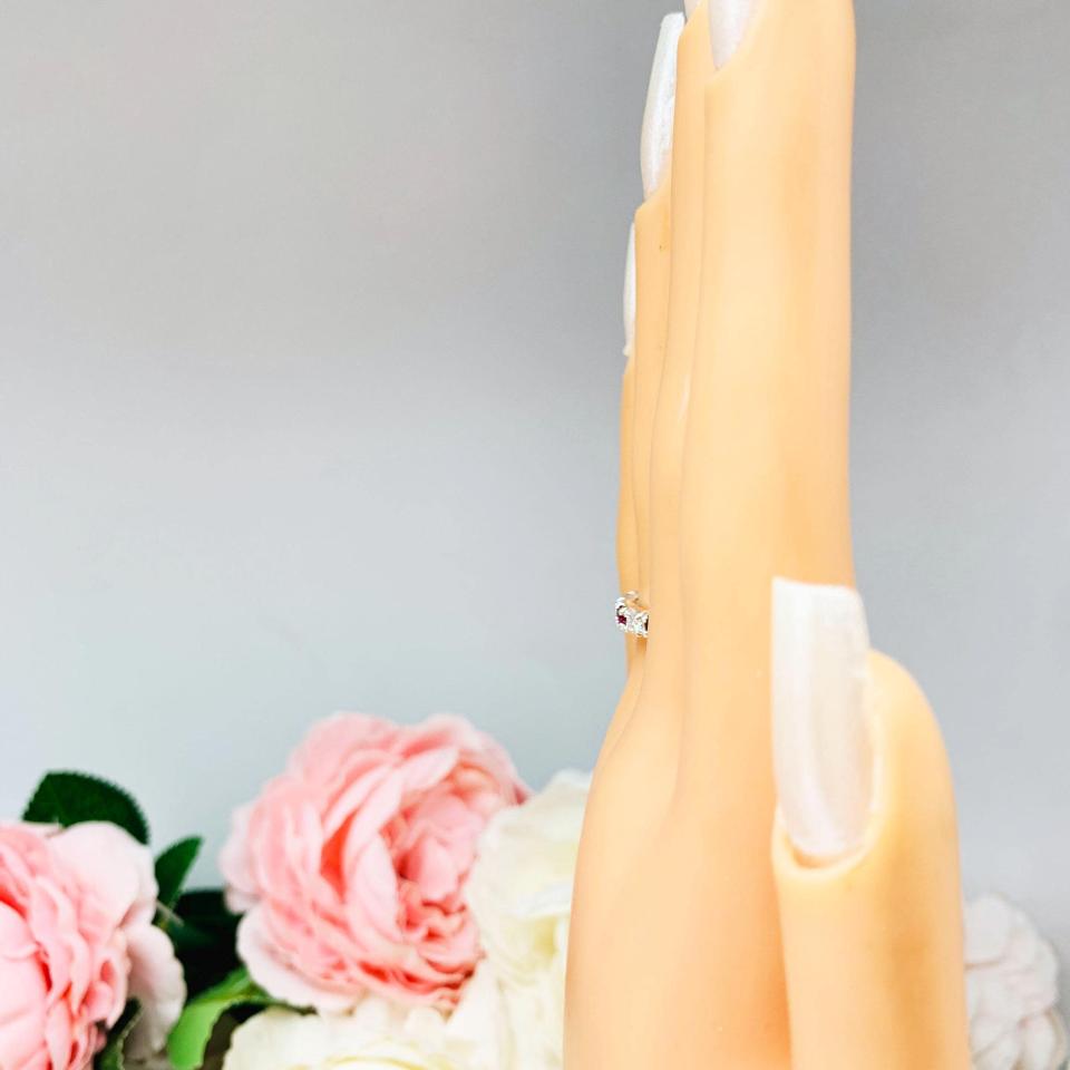 Mannequin hand with long white shimmery nails, displayed against a soft white-gray background with pink and white floral accents. The hand is wearing a sterling silver 5-stone band featuring alternating round lab rubies and moissanites. The band has a classic design with the gemstones set in a row with three rubies and two moissanites.