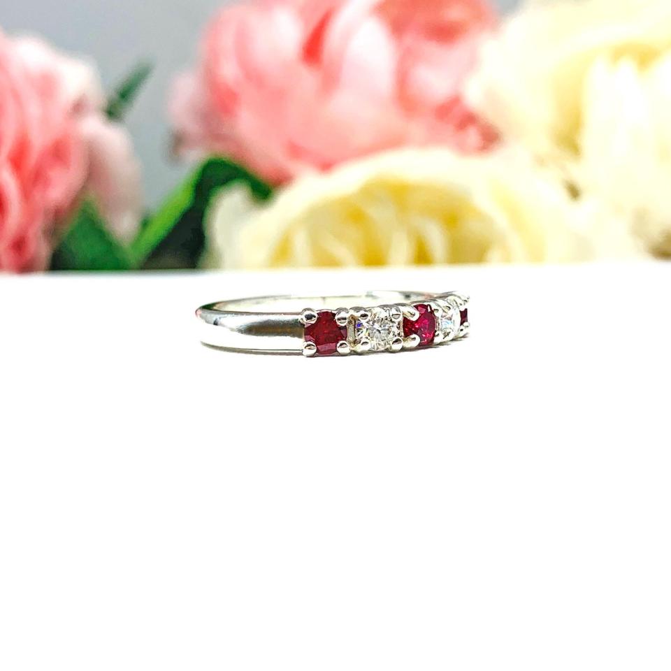 A sterling silver 5-stone band featuring alternating round lab rubies and moissanites. The band has a classic design with the gemstones set in a row with three rubies and two moissanites. Displayed on a white surface with a pastel floral background.