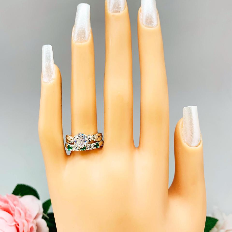 Mannequin hand with long white shimmery nails, displayed against a soft white-gray background with pink and white floral accents. The hand is wearing a sterling silver  five-stone wedding band with three 3mm round lab-created emeralds and two 3mm round DEF moissanites. Shown with a round moissanite solitaire leaf design ring (not included).