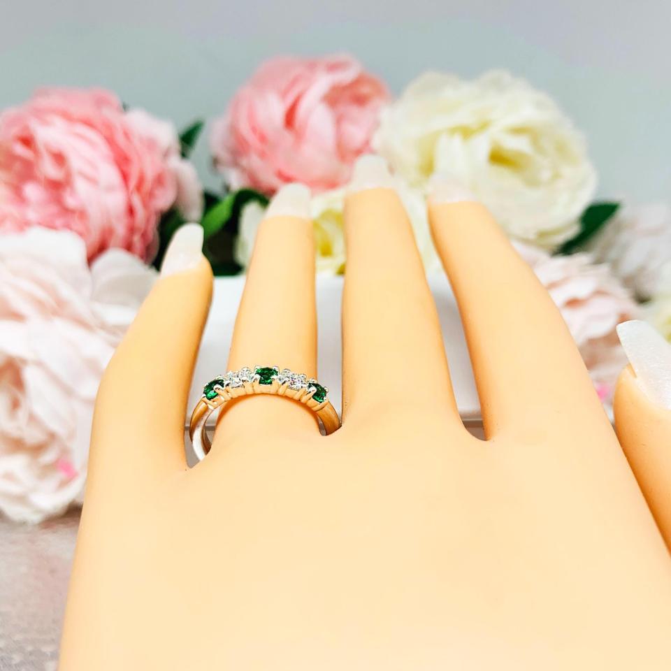Mannequin hand with long white shimmery nails, displayed against a soft white-gray background with pink and white floral accents. The hand is wearing a sterling silver  five-stone wedding band with three 3mm round lab-created emeralds and two 3mm round DEF moissanites.