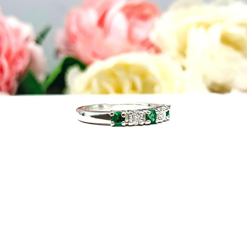 Sterling silver five-stone wedding band with three 3mm round lab-created emeralds and two 3mm round DEF moissanites, shown against a soft floral background.