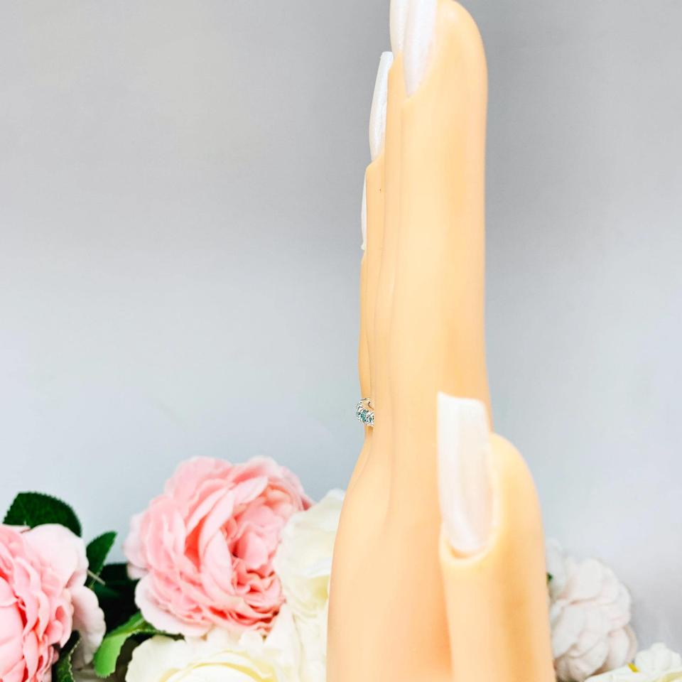 Mannequin hand with long white shimmery nails, displayed against a soft white-gray background with pink and white floral accents. The hand is wearing a sterling silver 5-stone band set with 3mm round sparkling aquamarine blue moissanites.