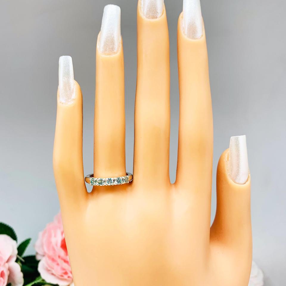 Mannequin hand with long white shimmery nails, displayed against a soft white-gray background with pink and white floral accents. The hand is wearing a sterling silver 5-stone band set with 3mm round sparkling aquamarine blue moissanites.