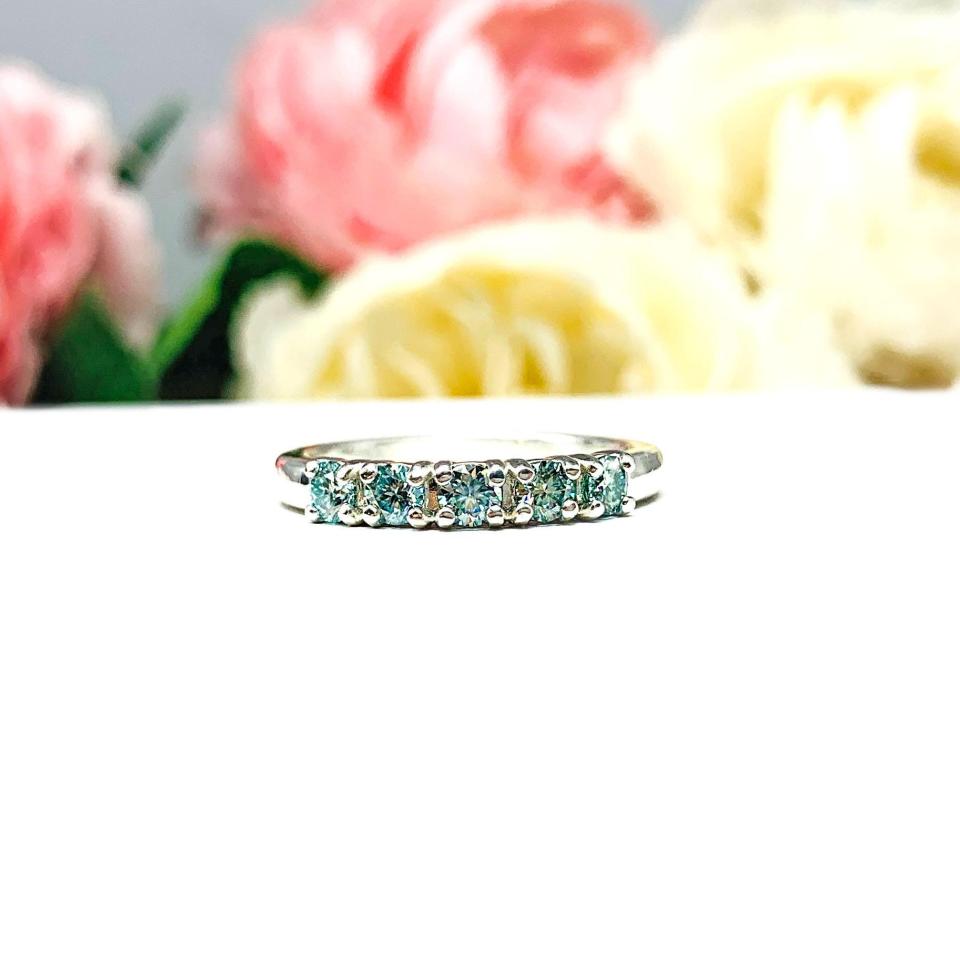 A sterling silver 5-stone band set with 3mm round sparkling aquamarine blue moissanites. Displayed on a white surface with a floral background.