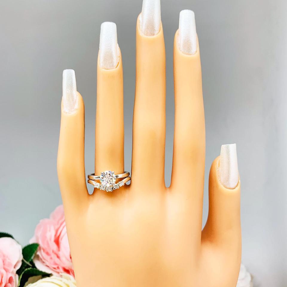 Mannequin hand with long white shimmery nails, displayed against a soft white-gray background with pink and white floral accents. The hand is wearing a sterling silver 5-stone curved wedding band featuring 2mm round precision faceted moissanite stones in prong settings. Shown with a round moissanite solitiare with (not included).