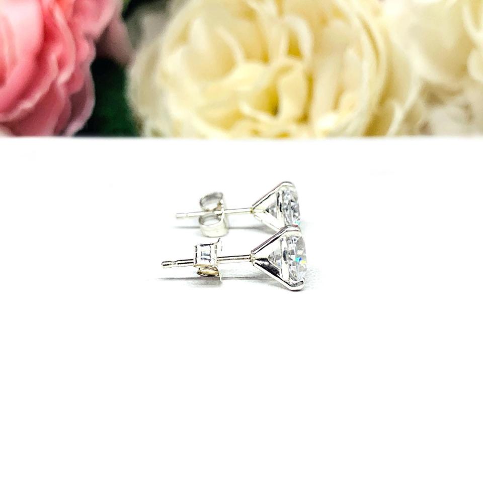 A pair of sterling silver 3-prong martini stud earrings with 6mm colorless old European cut cubic zirconias. The earrings are resting on a white surface with a background of pink and white flowers.