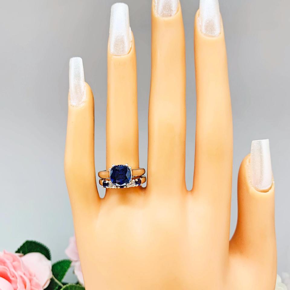 Mannequin hand with long white shimmery nails, displayed against a soft white-gray background with pink and white floral accents. The hand is wearing a sterling silver 5-stone band featuring alternating round lab blue sapphires and moissanites. The band has a classic design with the gemstones set in a row with three sapphires and two moissanites. It is featured here with a cushion-cut lab blue sapphire ring (not included).