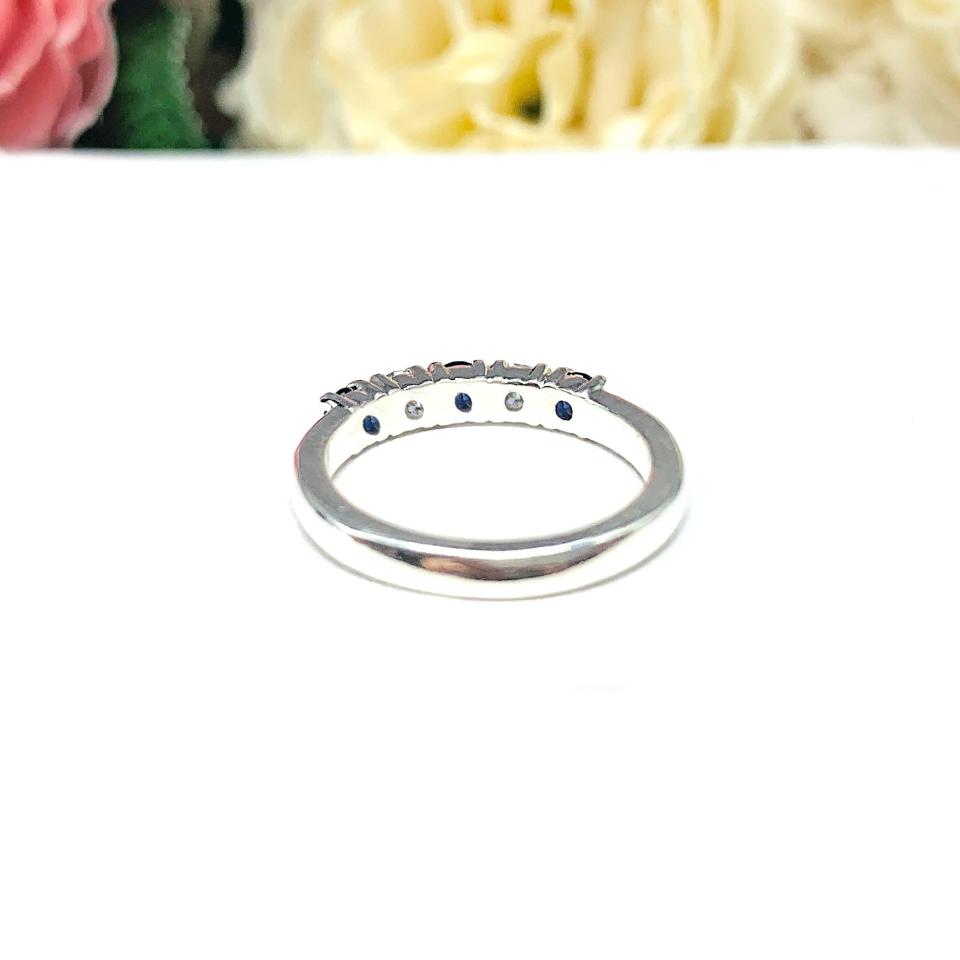A sterling silver 5-stone band featuring alternating round lab blue sapphires and moissanites. The band has a classic design with the gemstones set in a row with three sapphires and two moissanites. Displayed on a white surface with a pastel floral background.