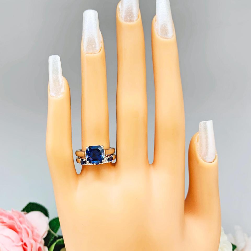 Mannequin hand with long white shimmery nails, displayed against a soft white-gray background with pink and white floral accents. The hand is wearing a sterling silver 5-stone band featuring alternating round lab blue sapphires and moissanites. The band has a classic design with the gemstones set in a row with three sapphires and two moissanites. It is featured here with an asscher-cut lab blue sapphire ring (not included).